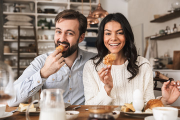 Image of young couple eating together at table while having breakfast in kitchen at home