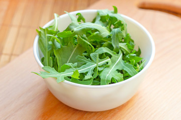 Fresh healthy green arugula leaves in a white bowl on light wooden background.