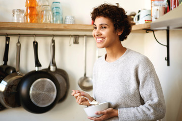smiling african american woman standing in kitchen eating breakfast cereal