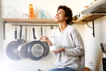 Poster happy young woman eating cereal from bowl in kitchen at home © mimagephotos