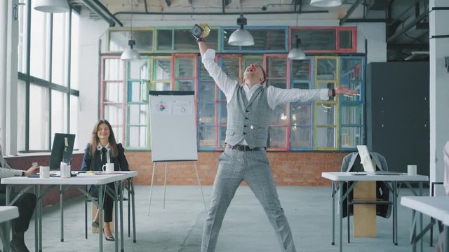 A young businessman enters the office with a reward in his hands. Colleagues join, dance with him and blow up flappers with confetti. Coworking in loft style. Office life. Winners