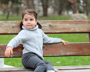 beautiful little girl with ponytails and in a sweater sitting on a bench in the park and smiling