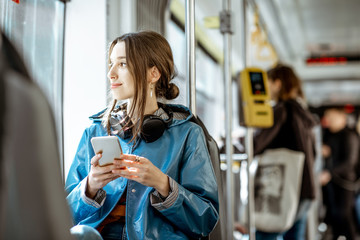 Young stylish woman using public transport, sitting with phone and headphones in the modern tram - obrazy, fototapety, plakaty