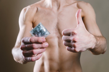 To the waist a naked, attractive man in black jeans holds a condom in his hands, and with his other hand he shows his finger upwards,