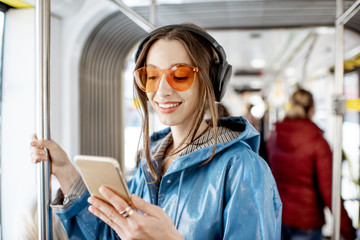 Young stylish woman using public transport, standing with headphones and smartphone while moving in...