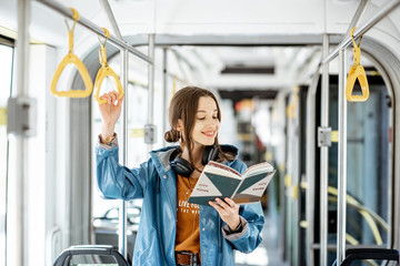 Young woman reading book while standing in the modern tram, happy passenger moving by comfortable...