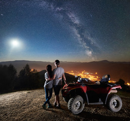 Rear view of happy young couple with atv quad motorbike on the top of mountain, boy and girl embracing, enjoying view of night sky full of stars, full moon, Milky way, luminous town on background © anatoliy_gleb