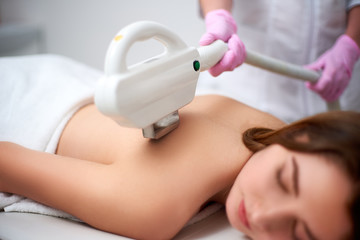 Young pretty smiling woman back laser epilation in beauty salon. Cosmetologist doing depilation...