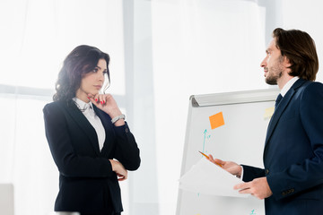 handsome man holding pencil while standing near white board and looking at pensive woman