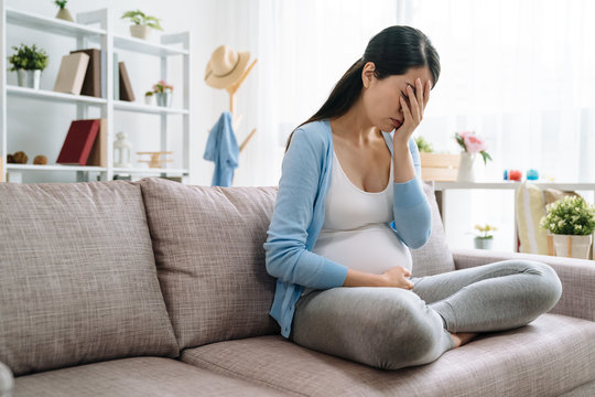 Sad And Stressed Pregnant Woman. Asian Young Female Lady At Home Depressed Not Ready To Be Mom Sitting On Couch Sofa At Home. Beautiful Upset Girl Cover Face By Hands Having Problem Parenthood