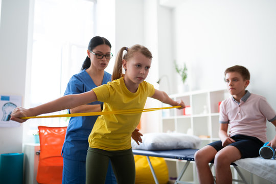 Attentive long-haired Asian therapist changing posture of little girl