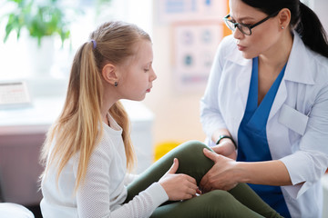 Caring dark-haired doctor in white robe checking state of hurting knee
