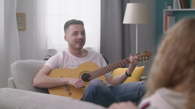 Panning of man plays the guitar for his friends in a cozy living room.