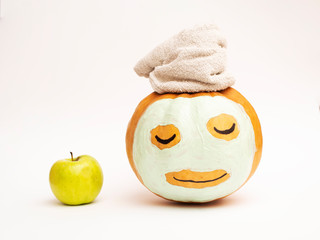 An apple and a pumpkin in the form of a face with a cosmetic mask and a towel on top