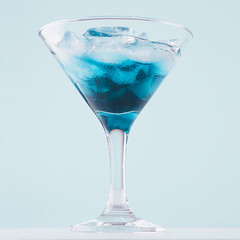 Summer refreshing blue drink with curacao, ice cubes in misted wineglass on soft light white wood board and pastel green interior, square.