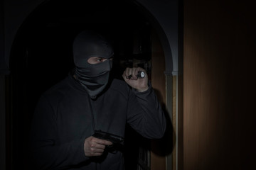 thief with a flashlight and a pistol in a mask with a slit for the eyes looking for prey in the apartment