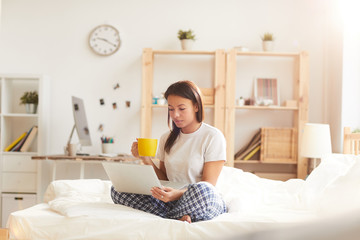 Obraz na płótnie Canvas Portrait of young Mixed-Race woman using laptop sitting on bed lit by sunlight, copy space