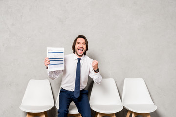 happy man in formal wear holding resume and gesturing in office