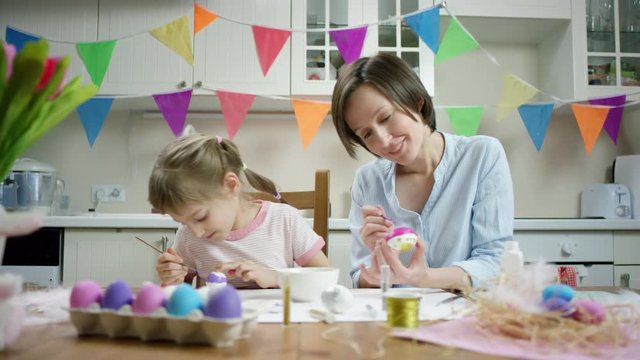 mother and daughter sitting at the table and crafting nest with colored eggs together and luaghing on the kitchen with overrun camera