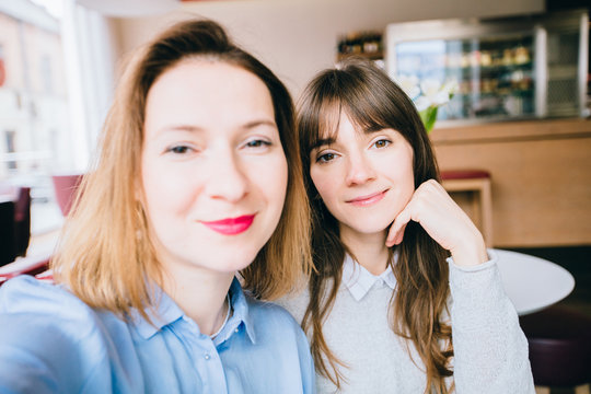 Two joyful attractive girls taking a selfie while sitting together at cafe looking at camera indoors. Adult middle age woman making selfie during lunch with sister.
