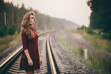 Beautiful girl with curly natural hair enjoy nature in forest on railway. Dreamer lady in burgundy dress walk on railroad. Female portrait of inspired girl on rails at dawn. Sun in hair in autumn.