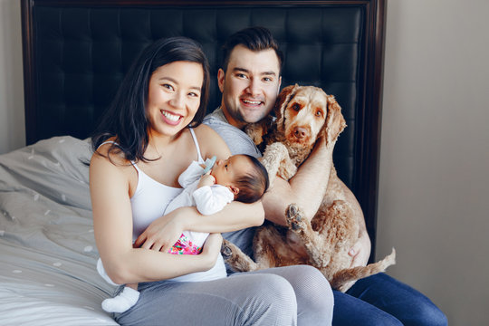 Smiling Chinese Asian Mother And Caucasian Father With Mixed Race Newborn Infant Baby Son Daughter And Large Pet Dog. Happy Family In Bedroom. Home Lifestyle Authentic Natural Moment.