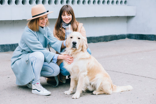 Two Happy Laughing Adult Twins Female Sisters With Running Away Dog. Training Golden Retriever, The Performance Of The Teams.