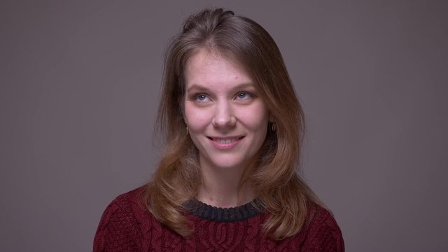 Pretty young female student watching smilingly into camera being shy on gray background.