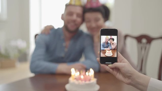 The female hand making a photo on the cellphone of happy smiling mature woman hugging with adult grandson, both in birthday caps. The cake is on the table in front of people. Birthday of grandmother