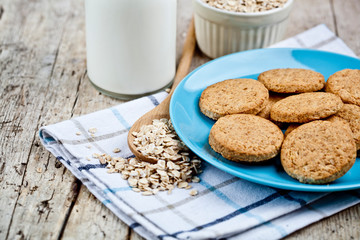 Fresh baked oat cookies on blue ceramic plate on linen napkin, bottle of milk and oak flakes on rustic wooden table.