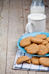 Fresh baked oat cookies on blue ceramic plate on linen napkin and cup of milk on rustic wooden table background.