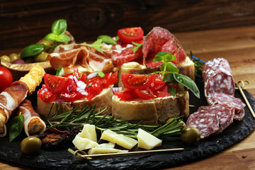 Traditional italian antipasto bruschetta appetizer with cherry tomatoes, cream cheese, basil leaves and balsamic vinegar on cutting board with prosciutto, salami, cheese