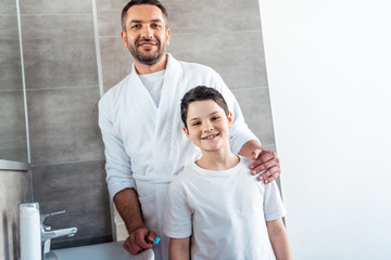 Fototapeta premium smiling father and son in pajamas looking at camera in bathroom