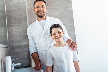 smiling father and son in pajamas looking at camera in bathroom