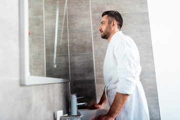 Fototapeta na wymiar handsome man in white bathrobe looking at mirror in bathroom during morning routine