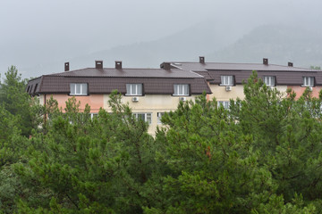 Fototapeta premium Pine trees and a residential building with a wet roof after the rain against the backdrop of mountains in the fog. Gelendzhik, Russia