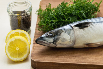 Lemon and black pepper near brown wooden cutting board with one raw mackerel and bunch of fresh...