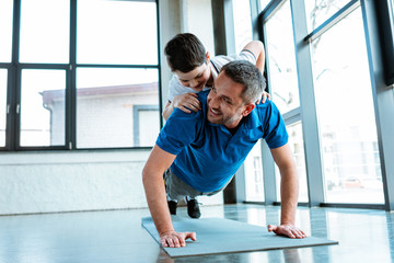 Happy father doing push up exercise with son on back at gym with copy space