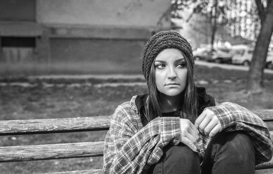 Homeless girl, Young red hair girl sitting alone outdoors on the wooden bench on the street with hat and shirt feeling anxious and depressed after became a homeless person black and white moody image