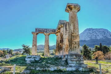 Columns in ruins of Temple of Apollo with Acrocorinth the acropolis of ancient Corinth - a monolithic rock overseeing the ancient city of Corinth Greece in the background and a tourist taking a phone 