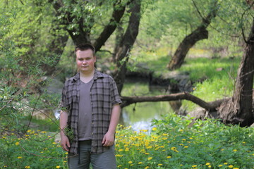 Young plump man stands with his back to the river. In the park among the bright green foliage.