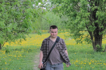 A young well-fed man walks through a spring meadow with bright green young grass, trees and flowering dandelions.