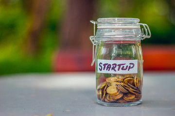 Closed small glass jar with coins in it and a label with the word startup written on it on a table with a green blurry background – Concept image for investment, a new start or raising founds