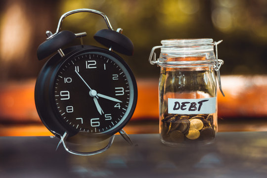 Glass jar with golden coins and a label with the word debt on it sitting next to a black alarm clock – Concept image for planning money and saving