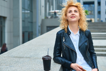 curly blonde young woman with a cityscape in the background holding a paper glass and a mobile phone