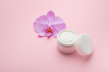 Mockup cosmetic cream with herbal flowers for face, skin and body care hygiene moisture lotion wellness therapy mask in plastic jar on pink background with orchid flower and copy space.