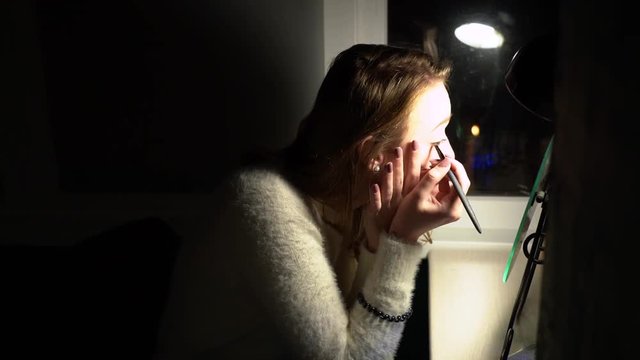 A pretty blond girl with acne on face in white pullover with earrings sits under black lamp, looks into a mirror on a wooden table and makeup her eyes with a thin brush at home against a dark window.