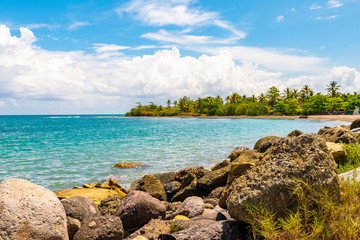 Obraz premium Coastal ocean view of endless seas on tropical Caribbean island. Large rock boulders along coastline, white sand beach with fishing boat painted in Rasta colors and coconut palm trees in background.