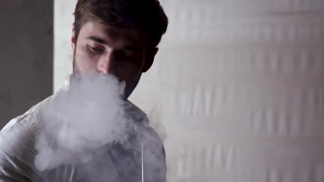 Brutal Caucasian bearded man with short brown hair in a white unbuttoned shirt smokes a hookah, exhales a cloud of smoke from his mouth and licks his lips at home. The concept of rest.