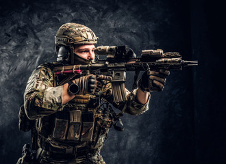 Close-up studio photo against a dark textured wall. The elite unit, special forces soldier in camouflage uniform holding an assault rifle with a laser sight and aims at the target
